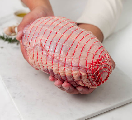 Raw boneless lamb leg roast wrapped in red netting, held over a white marble surface with rosemary and garlic in the background.