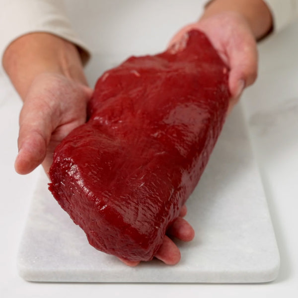 Hands holding a venison boneless short loin, placing it on a marble board, and slicing a piece off.