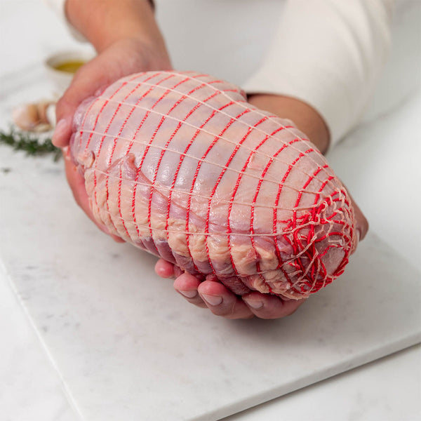 Raw lamb boneless leg roast wrapped in red netting, held over a white marble surface with rosemary and garlic in the background.