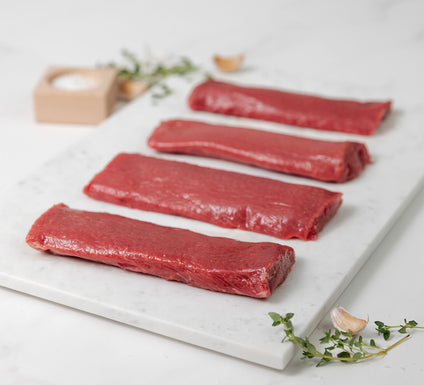 Four raw lamb loin fillets on a marble cutting board with herbs and salt in a wooden block in the background.