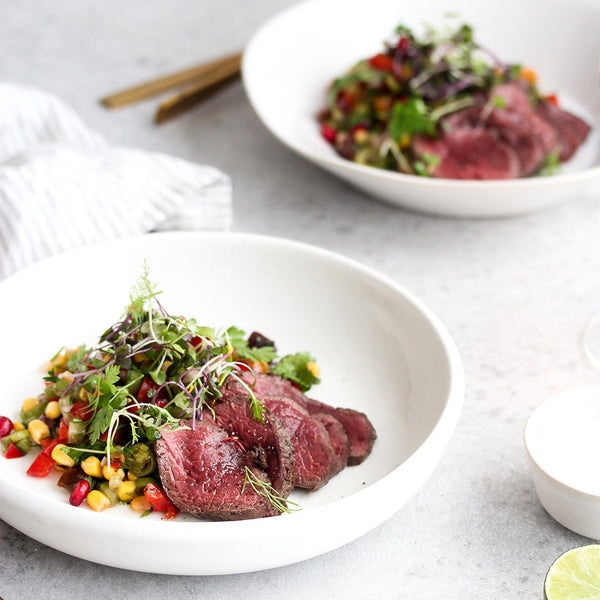 Two plates of venison slices with greens on a light gray surface.