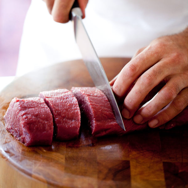 Person slicing raw venison short loin on a wooden cutting board with a knife.