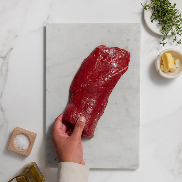 Raw venison short loin on a marble cutting board with a hand holding it, surrounded by butter, salt, olive oil and herbs.