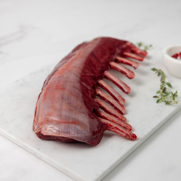 Raw rack of venison on a white marble cutting board with herbs and spices.