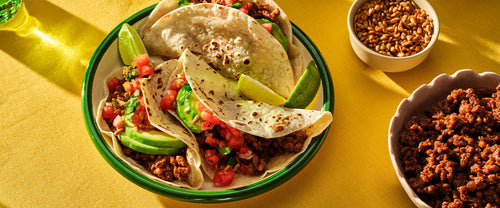 Three beef tacos with pico de gallo, avocado slices, and lime wedges on a plate, with bowls of seeds and extra beef on the side.