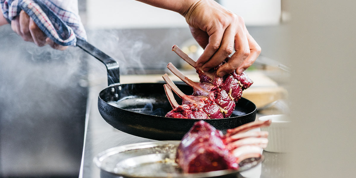 Person searing a venison frenched rack in a steaming cast iron skillet on a stovetop, with another rack resting on a tray in a professional kitchen.