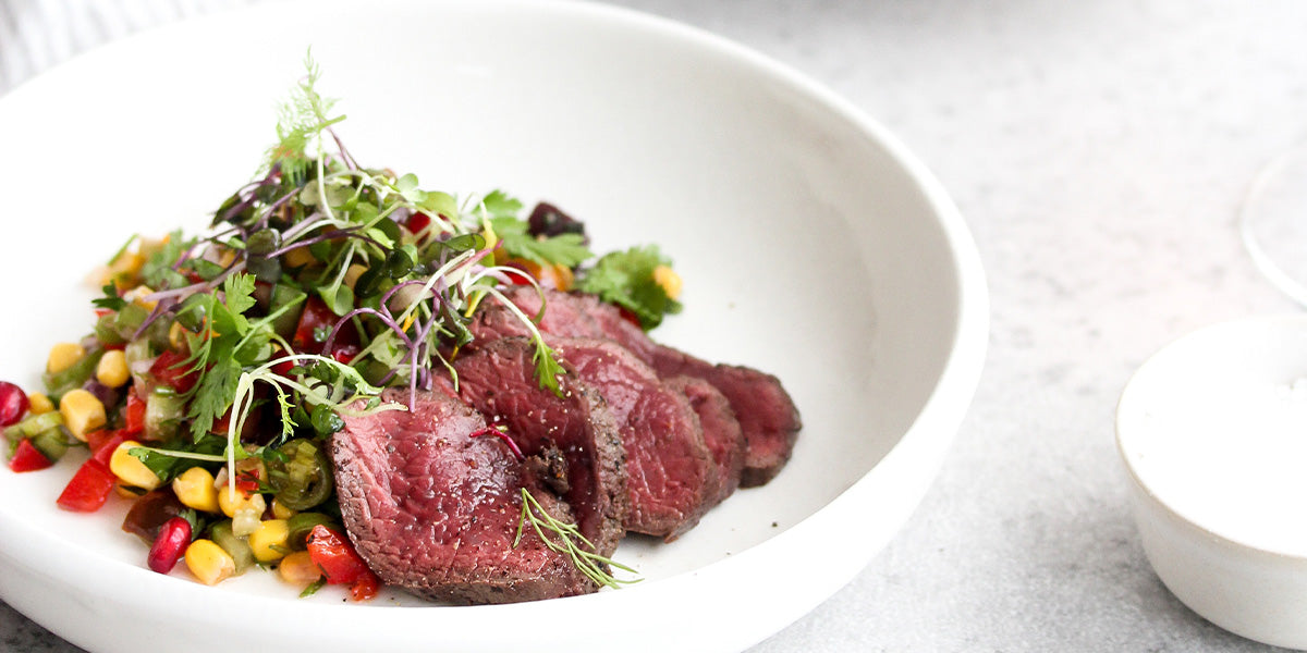 White bowl with medium-rare venison short loin slices topped with microgreens, served alongside a colorful vegetable medley.
