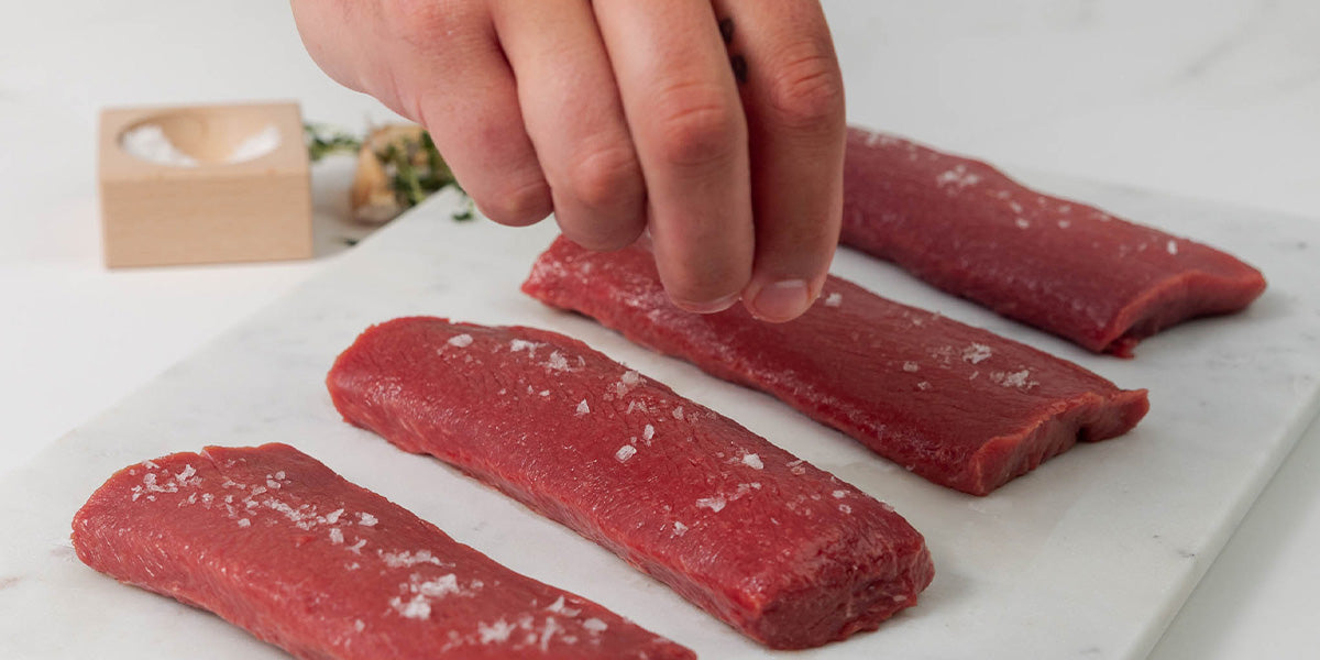 Hand seasoning 4 raw lamb loin filet cuts with salt on a white surface, showcasing premium meat preparation.