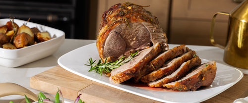 Sliced roasted boneless leg of lamb arranged on a white platter with fresh rosemary garnish.In the background, a dish of crispy roasted baby potatoes