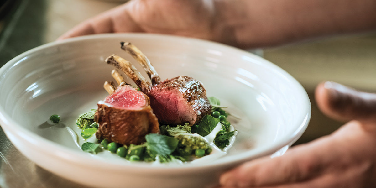 Medium-rare rack of lamb served over green vegetables in a white bowl, held by two hands.