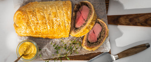 A gourmet venison Wellington featuring a perfectly cooked short loin wrapped in golden, flaky puff pastry, served in thick slices to reveal the tender, medium-rare meat inside.
