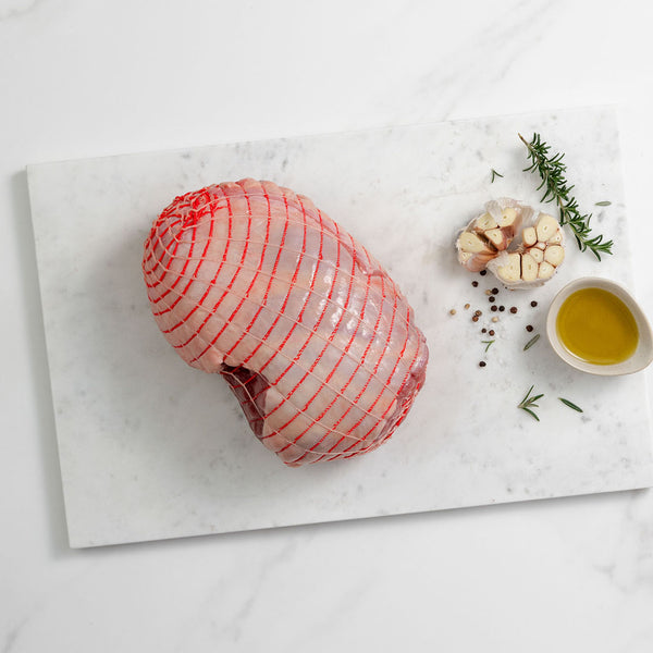 Raw boneless lamb leg wrapped in red netting on a white marble surface, with garlic, herbs, and a bowl of oil to the side—ideal for roasting or gourmet meal kits.