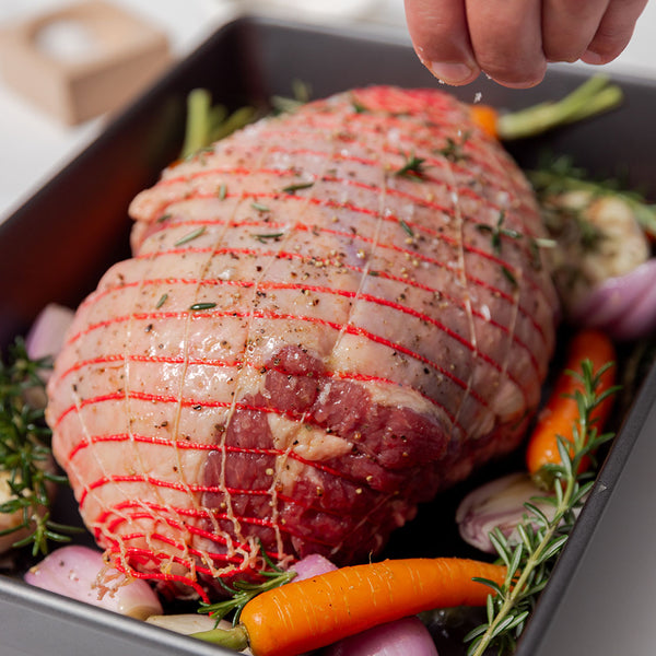 Raw Lamb Boneless Leg roast netted with red string in a baking tray, surrounded by carrots, onions, and rosemary—being seasoned with salt - ideal for oven-ready gourmet meals.
