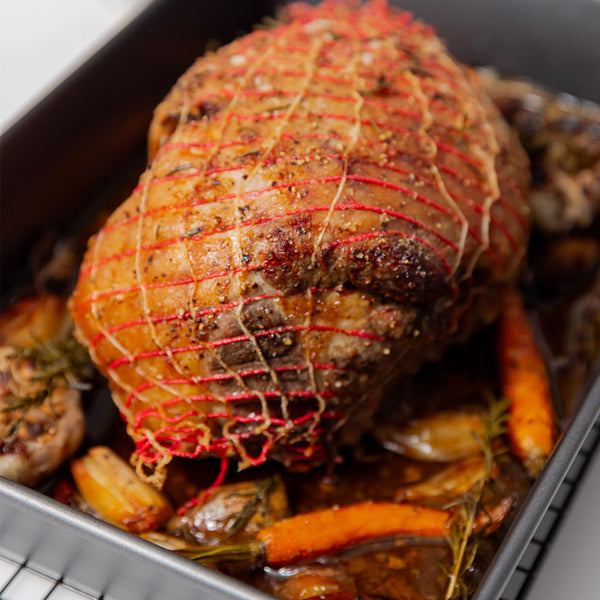 Cooked Lamb boneless leg roast in red netting, seasoned and surrounded by roasted carrots, garlic, and onions in a roasting pan—perfect for gourmet meal delivery or holiday dinners.
