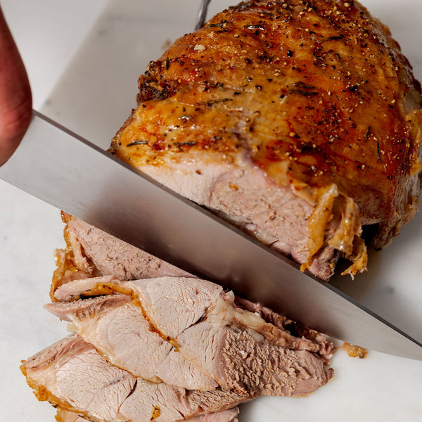 Carving a roasted bonless lamb leg roast with golden-brown crust on a white cutting board, showing juicy slices—perfect for premium meat lovers.