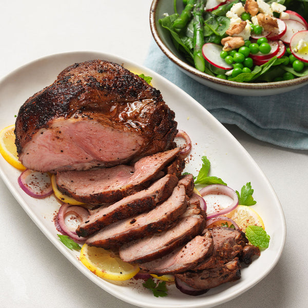 liced roasted lamb leg garnished with lemon, red onion, and herbs on a serving plate, with a spring salad of asparagus, radish, peas, walnuts, and feta in the background—ideal for entertaining or gourmet meal inspiration.
