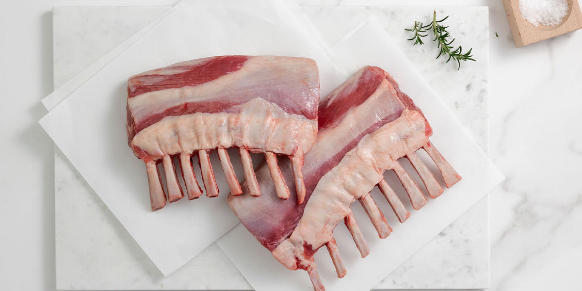 Raw lamb racks on a white marble surface with rosemary and salt.