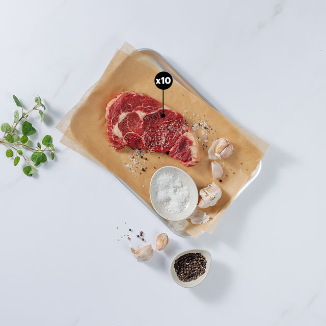 10oz Rib-eye Beef Steak sprinkle with salt and ground pepper placed on paper and tray showing x10 blurb with garlic cloves, herbs, pepper and salt in bowl at the side