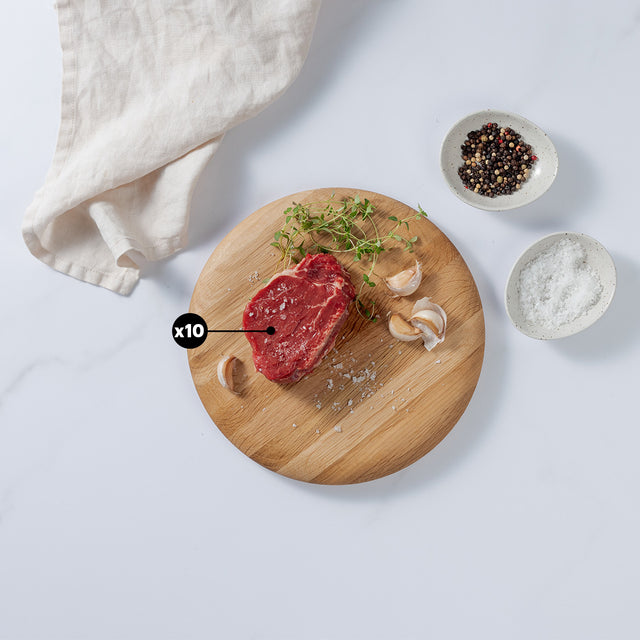 6oz Tenderloin Beef placed on round board showing x10 blurb with garlic cloves, herbs, and salt. Pepper and salt in bowl, kitchen towel at the side.