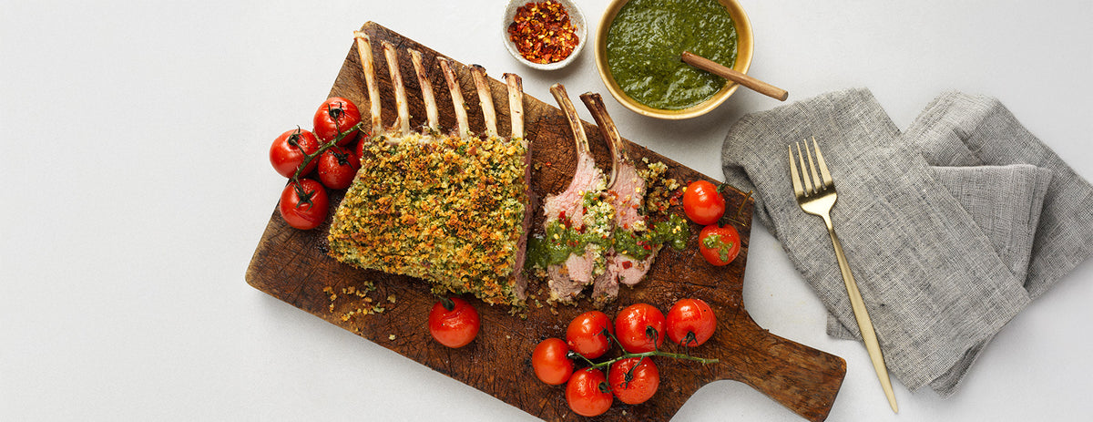 Plated dish of roasted rack of lamb with tomatoes on a wooden board, accompanied by a bowl of mint sauce, chili flakes and a fork.