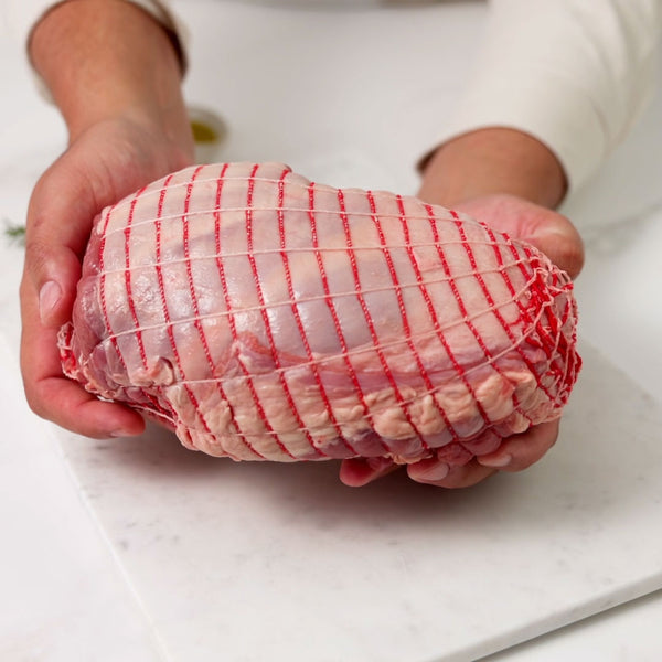 A pair of hands holding a raw, netted, boneless lamb leg, rotating it gently before placing it on a white marble board.