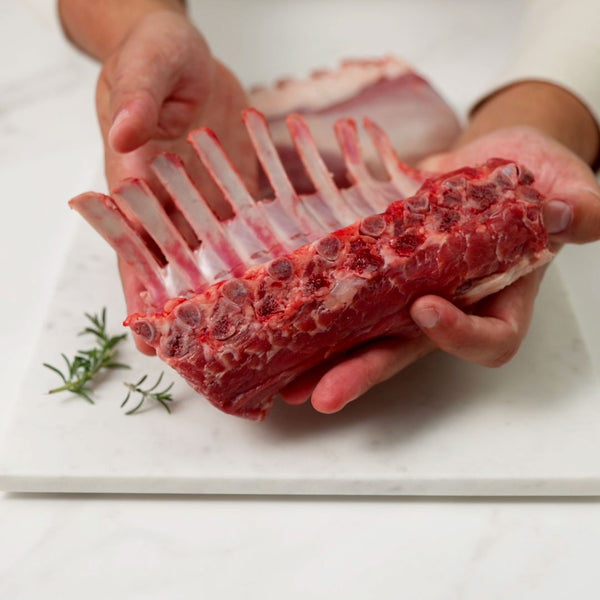 Holding a raw frenched lamb rack, gently rotating it before placing it on a white marble board, then slicing into individual chops.
