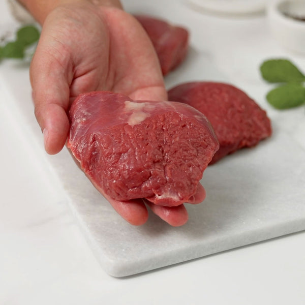 Holding a piece of raw lamb rump, gently rotating it before placing it on a white marble board alongside the other lamb rumps.