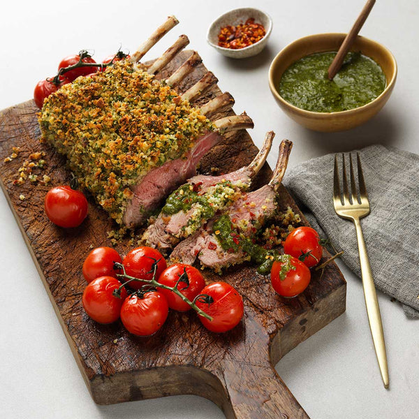 Roasted rack of lamb with herbs on a wooden cutting board with cherry tomatoes, accompanied by a bowl of green sauce.