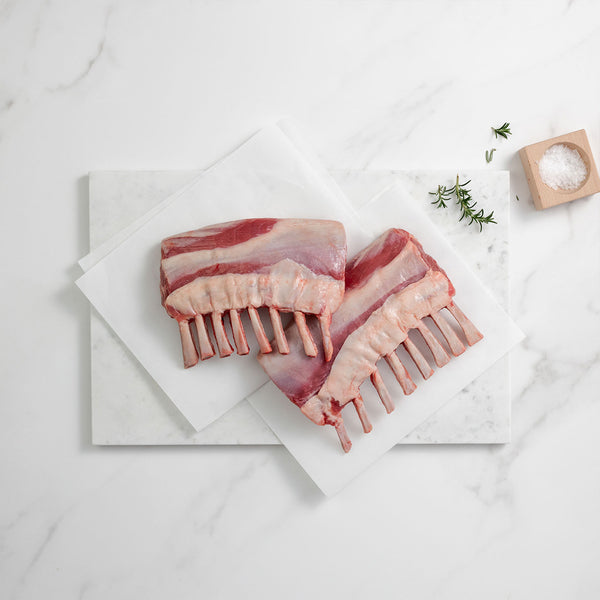 Raw lamb racks on a marble surface with a small container of salt and herbs.