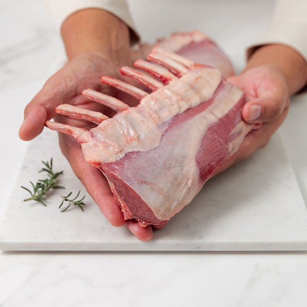Raw lamb rack held by hands on a white surface with rosemary sprigs.