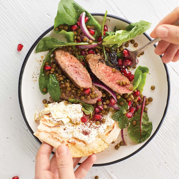 Salad with sliced lamb loin meat, greens, and pomegranate seeds on a white plate with a hand holding a fork.