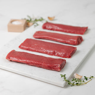 Four raw lamb loin fillets on a marble cutting board with herbs and salt in a wooden block in the background.
