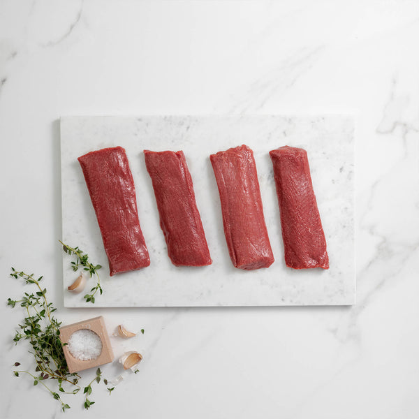 Four raw lamb loin fillets on a marble slab with herbs and salt on a light background
