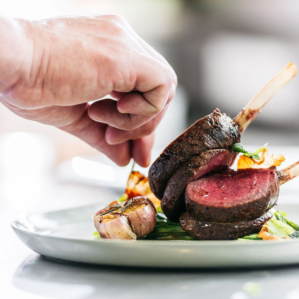 Close-up of a hand garnishing a dish with venison cooked ribs on a white plate.