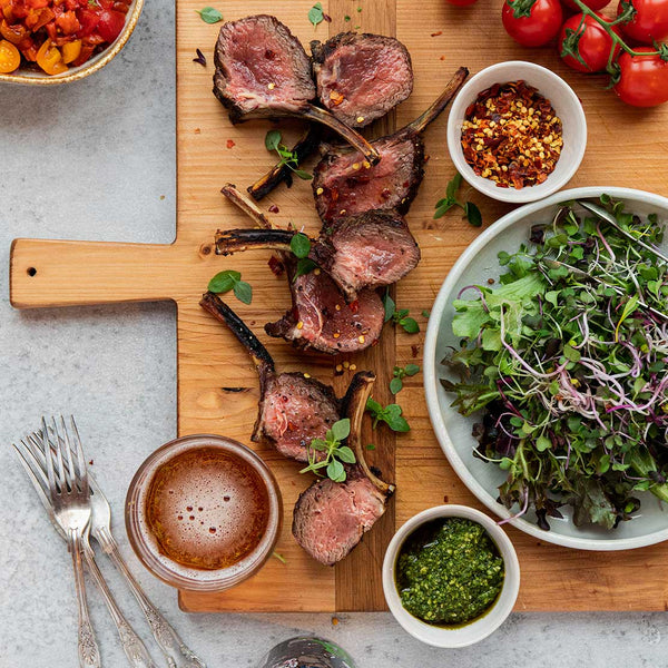 Wooden cutting board with cooked venison rib chops, side dishes, and utensils on a gray surface.