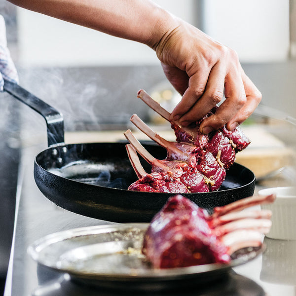 Person placing raw venison rack with 5 ribs into a frying pan on a stove.